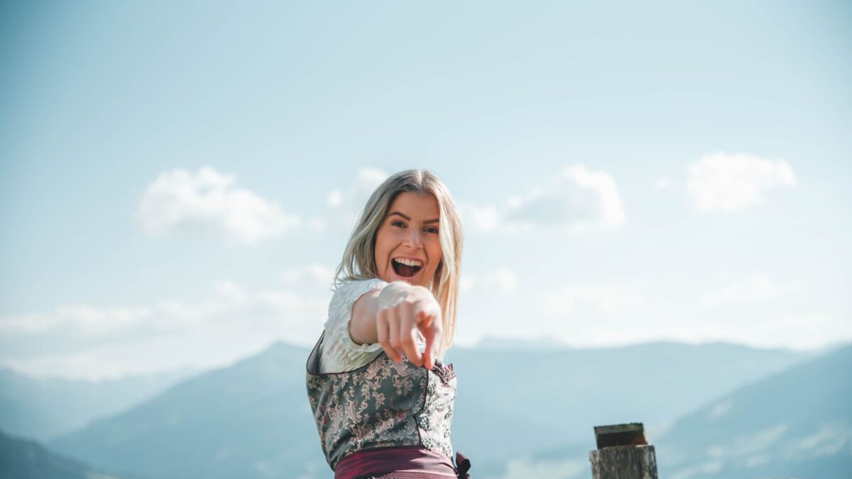Blond woman in a traditional dirndl pointing joyfully with mountains in the background.