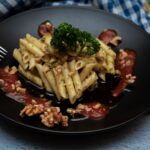 Penne pasta with walnuts and tomato sauce on a black plate, garnished with parsley.