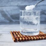 A spoonful of sugar being added to a glass of water on a wooden mat.