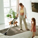 Pregnant mother cleaning carpet with vacuum as daughter and dog play indoors.