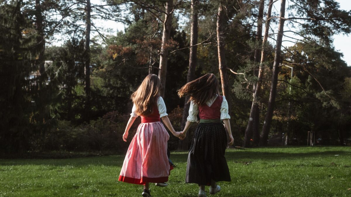 Two girls in traditional attire holding hands while walking in a lush green park on a sunny day.