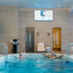 mediterana - Two adults enjoying leisure time at an indoor swimming pool with spa-like ambiance.